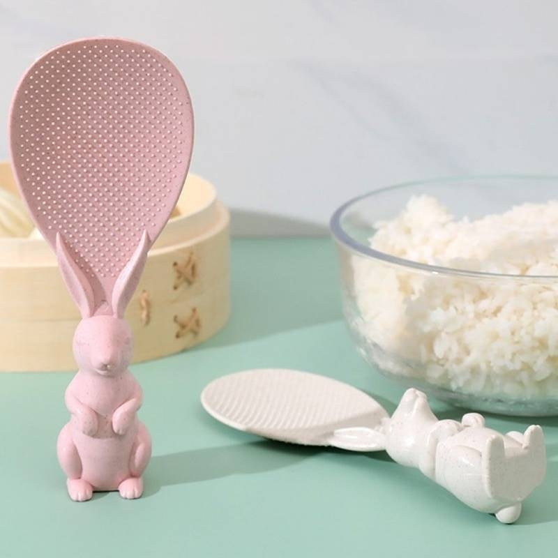 Pink rabbit-shaped rice paddle and white hippo-shaped spoon on a light green surface with a bowl of rice in the background.