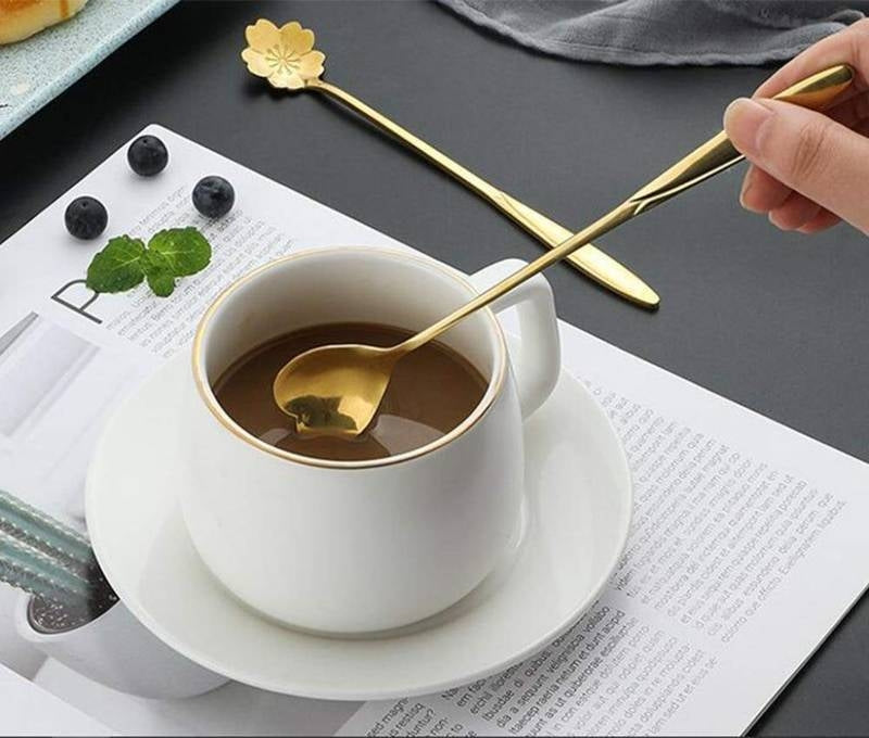White mug with gold spoon on a saucer, surrounded by blackberries and green leaves, on a newspaper.