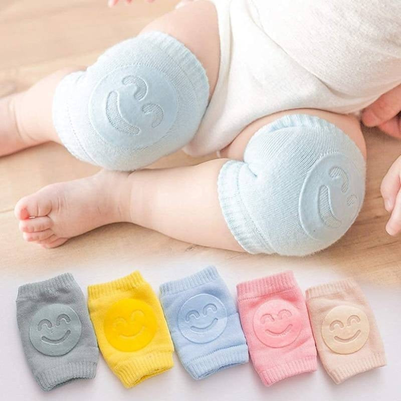 Baby wearing light blue knee pads with smiley face design on a wooden floor.